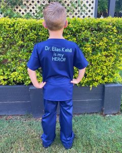 Photo of Korbin wearing hand made blue scrubs with back to camera with text saying Dr Elias Kehdi is my HERO!