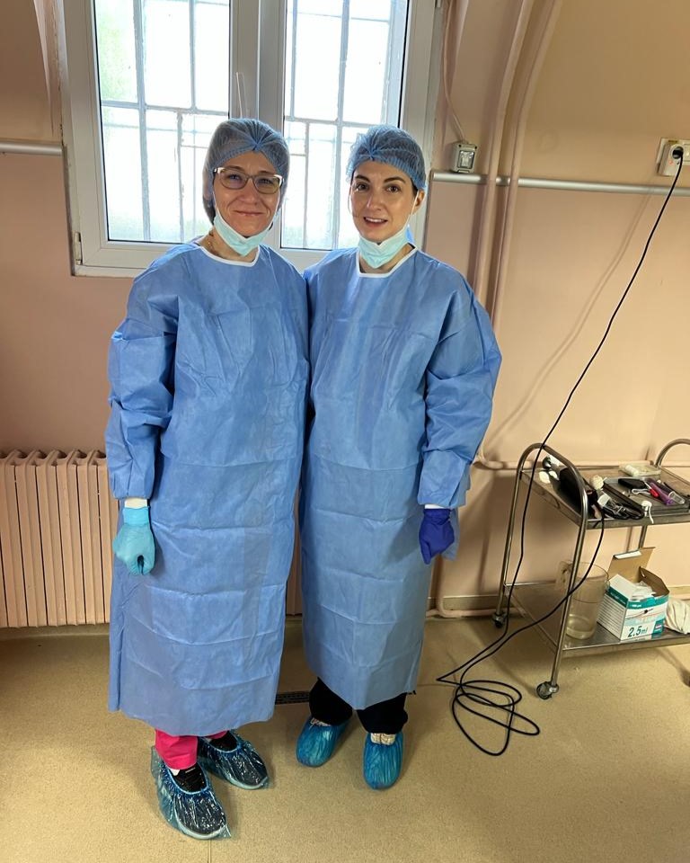 Two women in blue surgical gowns and gloves, wearing paper hair covers and disposable masks, lowered to show their faces, stand side by side in a room with apricot couloured walls. Beside them is a stainless steel trolley holding surgical equipment.
