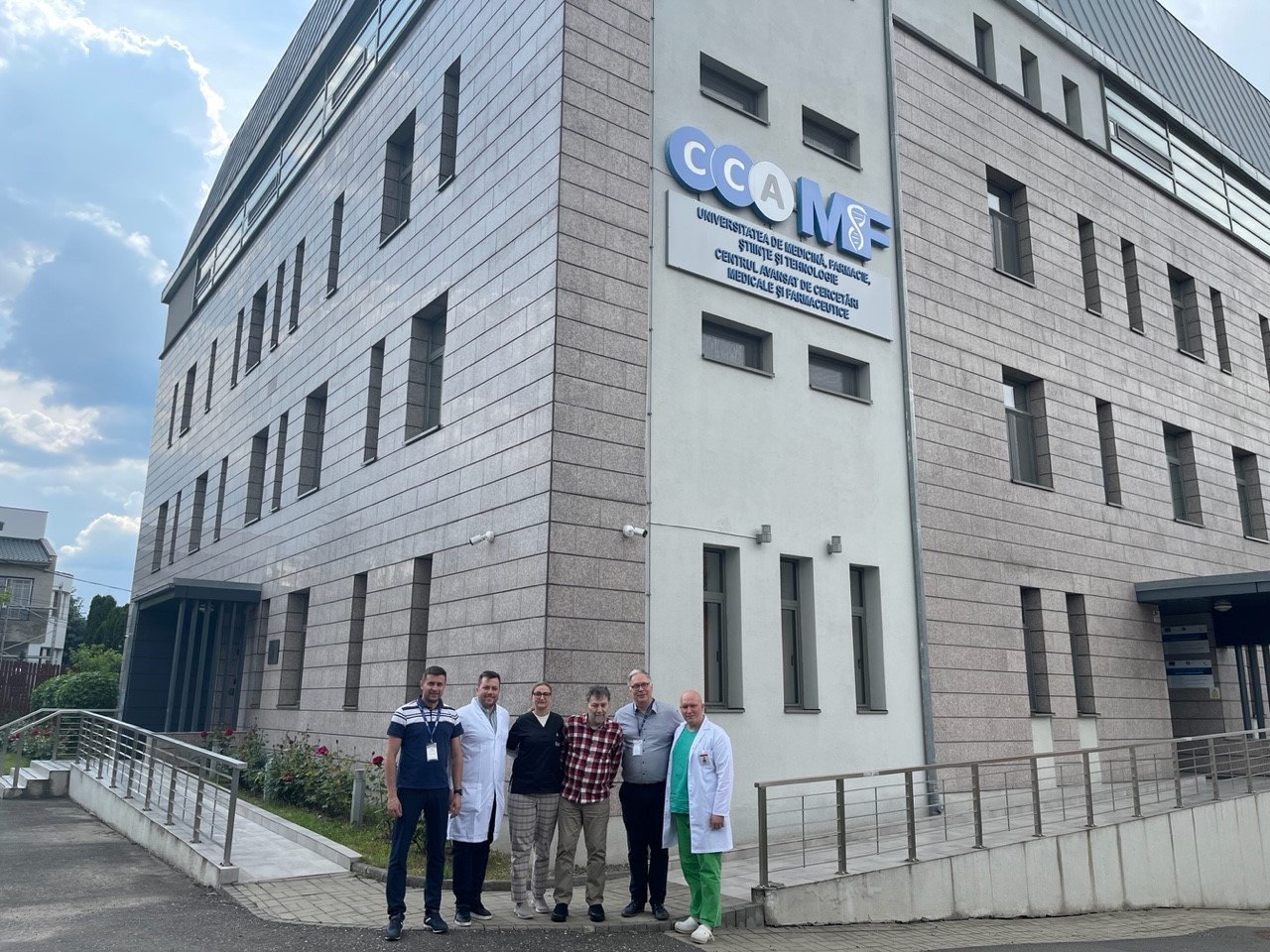A group of people, five men and one woman, stand shoulder to shoulder in front of a four storey brick building with a sign in Romanian, indicating the building is a centre for medical research.
