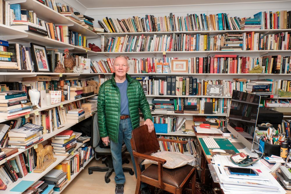 An older man with grey hair in a blue jumper, jeans and green puffer jacket stands behind a desk bearing a large computer monitor, books and papers. Behind him there are floor to ceiling shelves holding piles of books, framed photographs and mementos