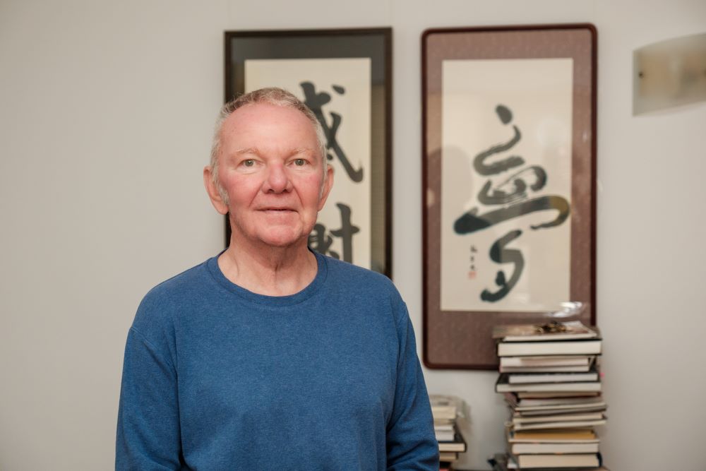 An older man with grey hair in a blue, round necked jumper stands in front of two framed calligraphy artworks and a table with piles of books on it