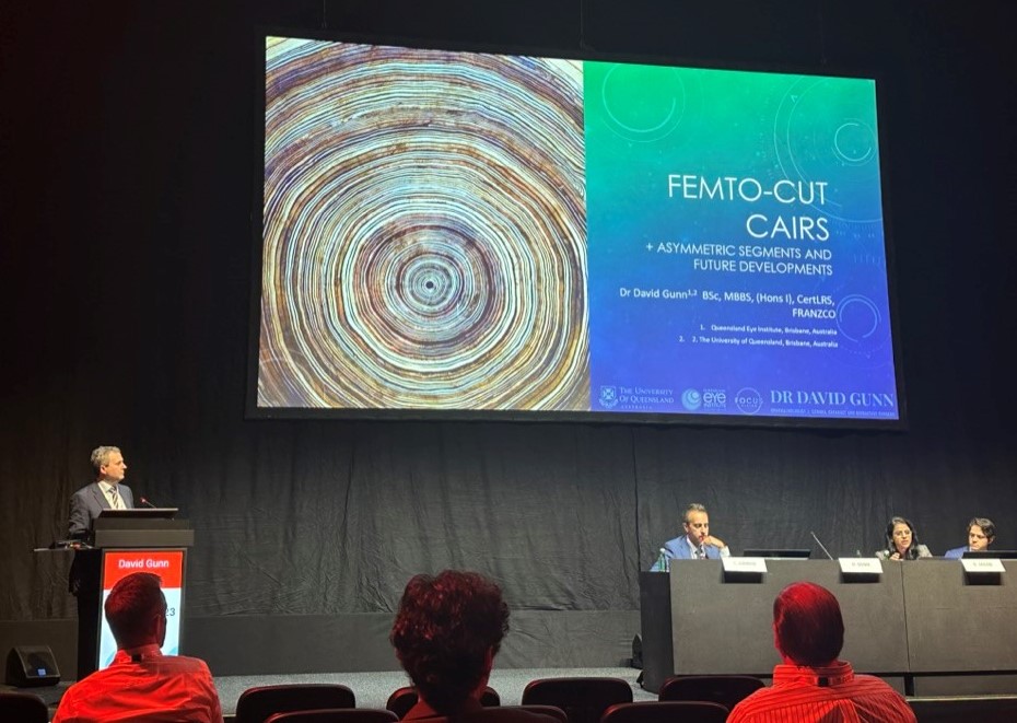 Dr David Gunn stands on a stage behind a lectern, speaking into a microphone, addressing an audience in an auditorium. There is a desk on the stage with three people seated behind it. On the wall behind the stage is a large screen with a projection of a slide which reads “FEMTO-CUT CAIRS – ASYMMETRIC SEGMENTS AND FUTURE DEVELOPMENTS”