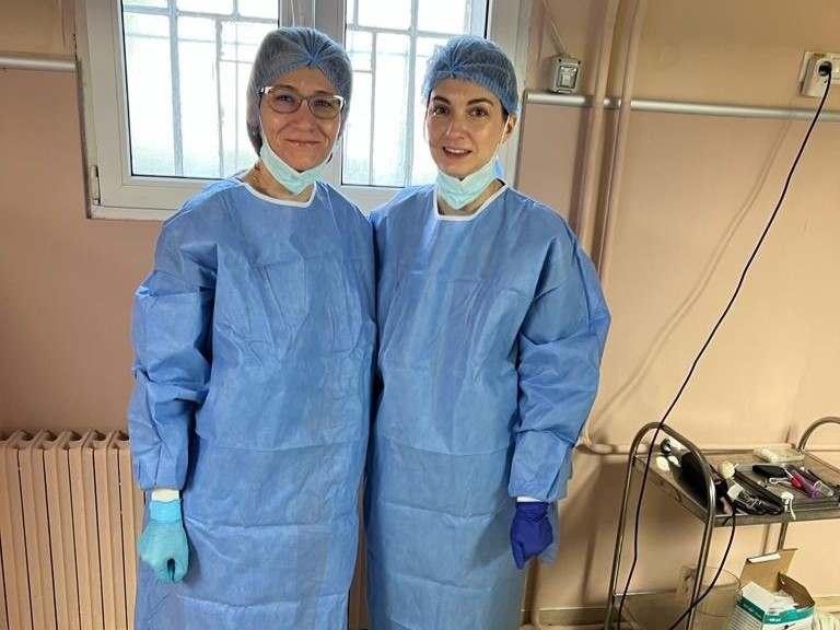Two women in blue surgical gowns and gloves, wearing paper hair covers and disposable masks, lowered to show their faces, stand side by side in a room with apricot coloured walls. Beside them is a stainless steel trolley holding surgical equipment.