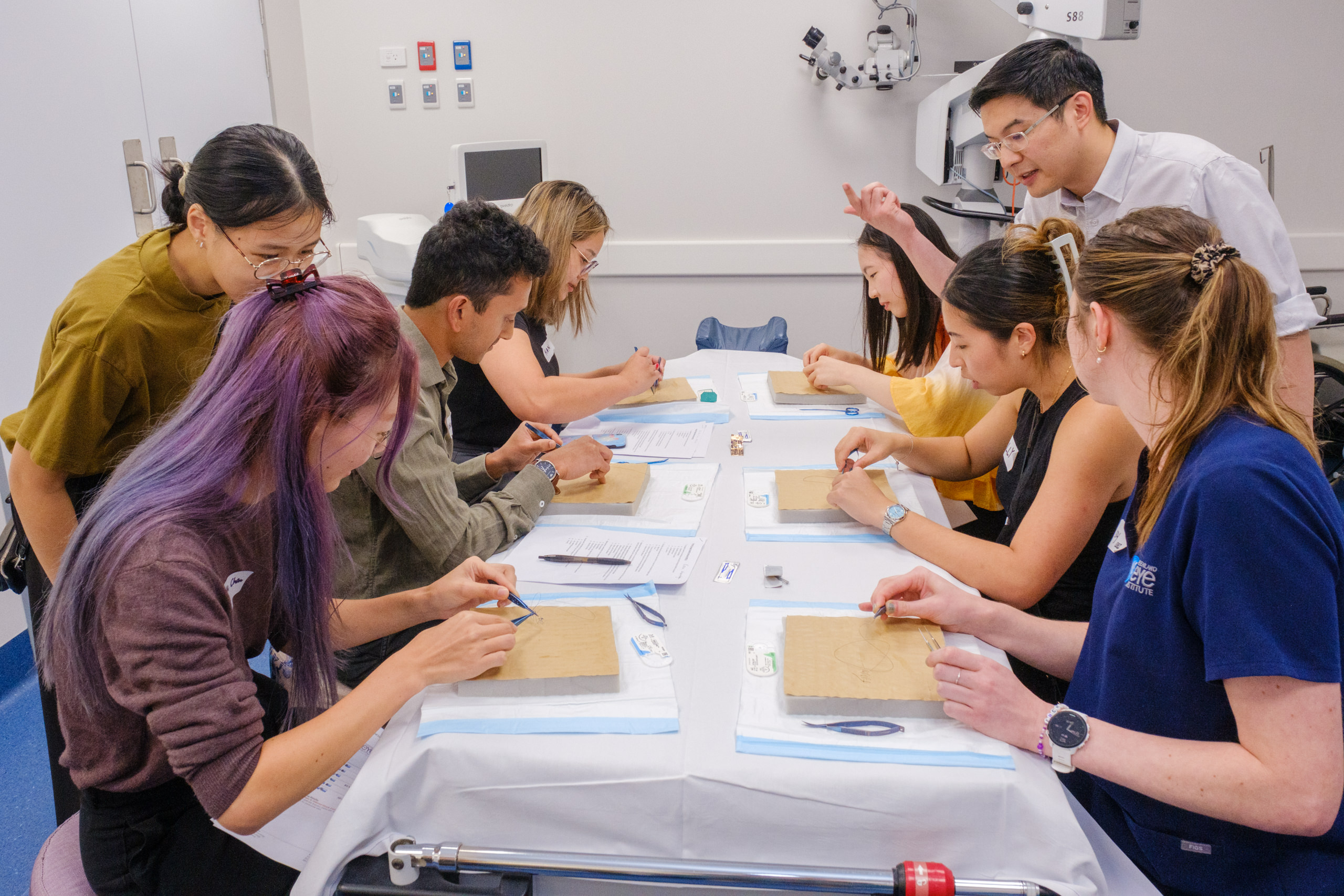 Seven people sit or stand around a table with square foam blocks in front of them, using surgical implements to practise suturing. A man with short dark hair and glasses lifts his hand to demonstrate the suturing technique.