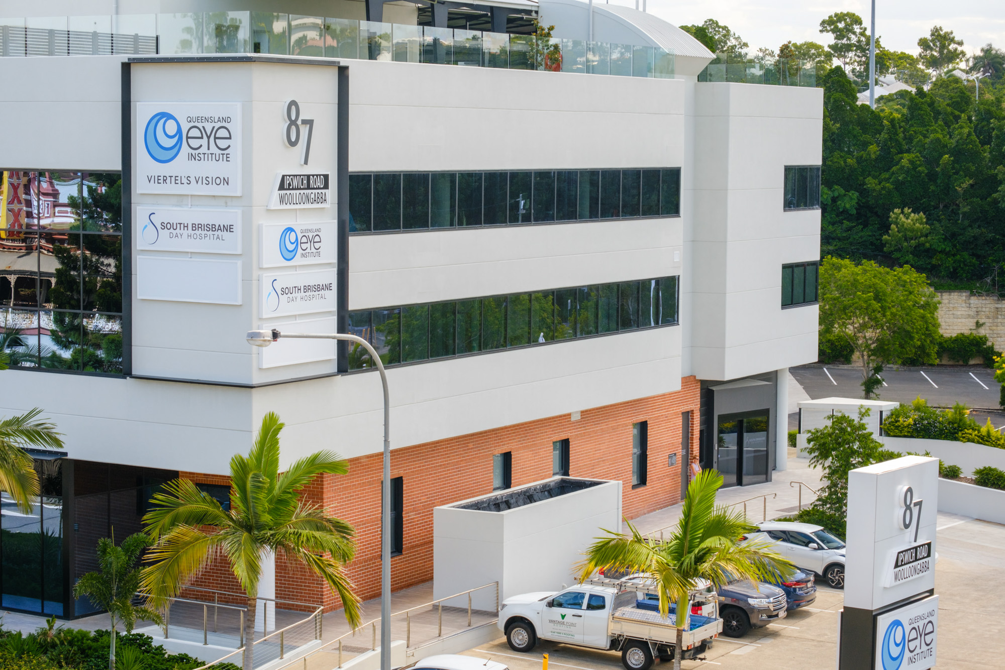 A four storey building with red brick on the ground floor and white cladding above with dark tinted windows. There are sings on the building reading "87 Ipswich Rd, Queensland Eye Institute, Viertel's Vision, South Brisbane Day Hospital"