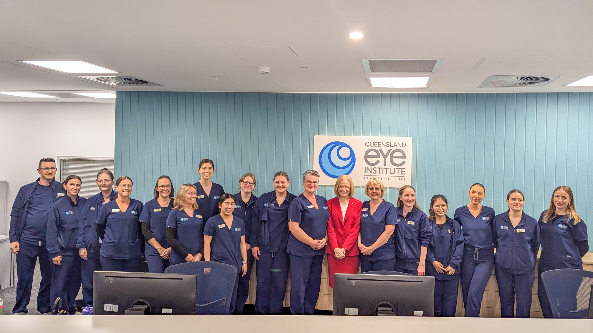 A woman in a red suit stands surrounded by a group of 17 men and women in blue medical scrubs in front of a wall with a sign reading "Queensland Eye Institute"