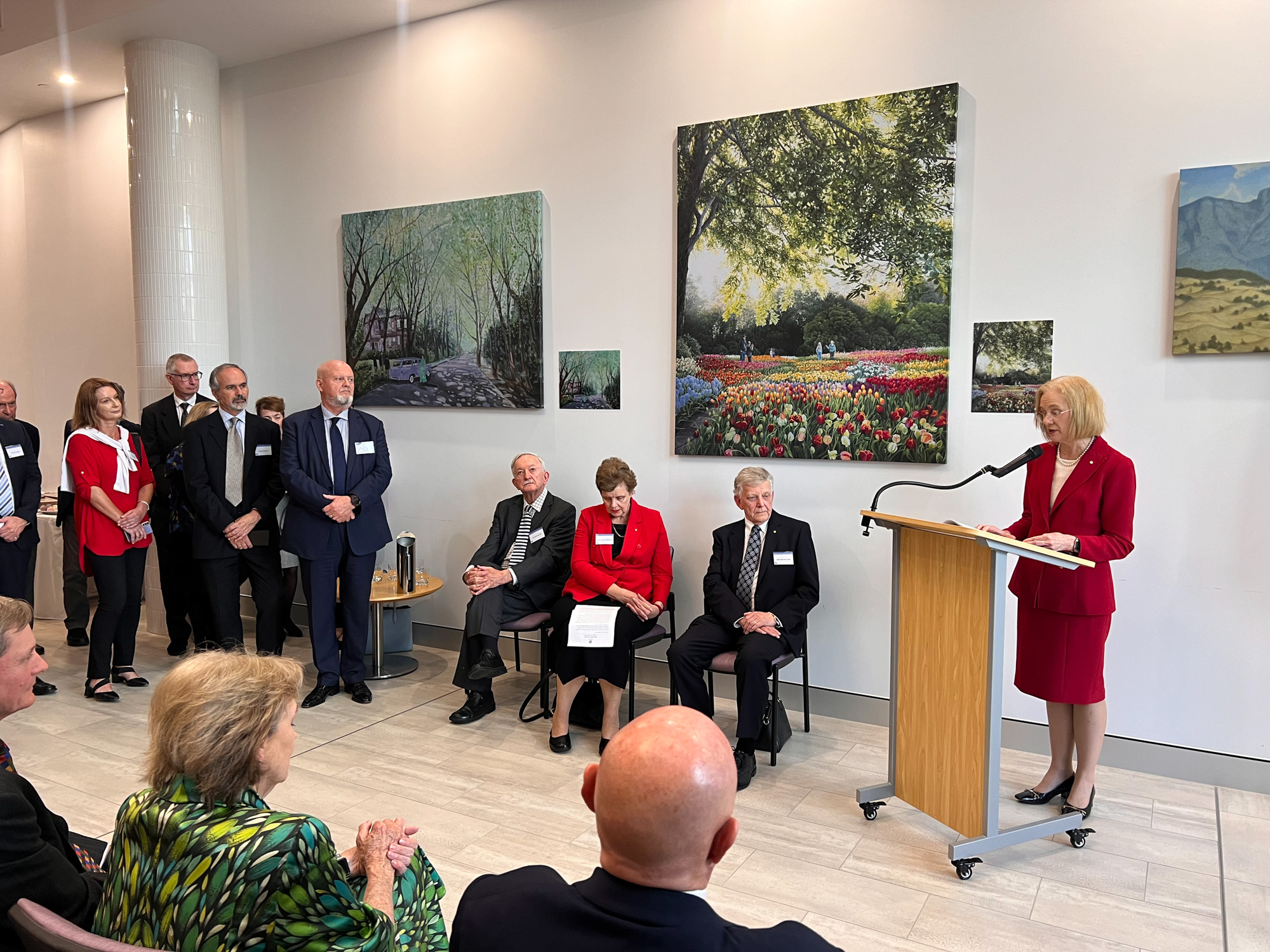 A woman in a red suit delivers a speech from behind a lecturn with an audience of men and women in suits looking on. There are large artworks on the wall behind the speaker.