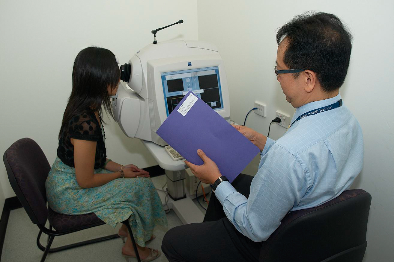 A man holding a folder looks at the screen of an eye examination screen while a woman looks into the eye piece on the machine