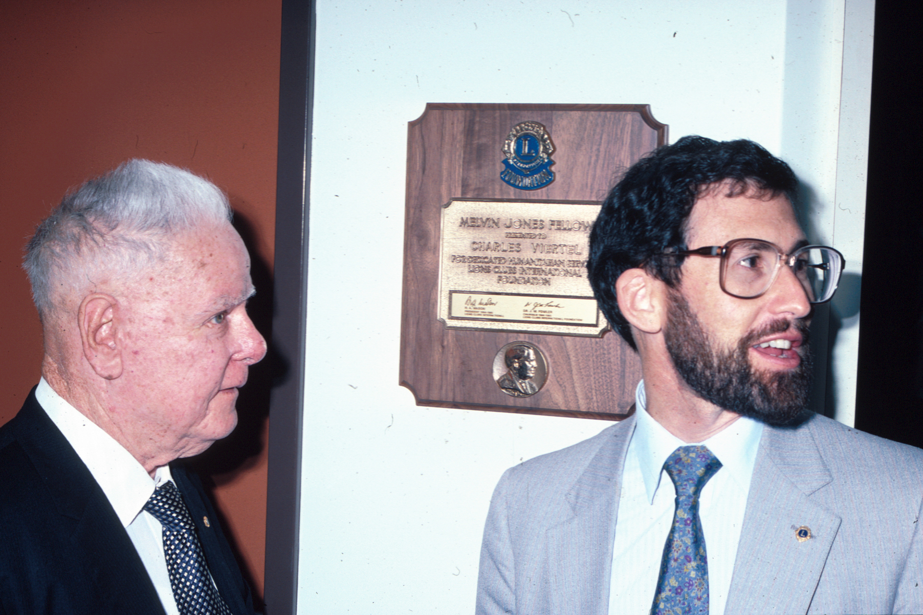 Close up of two men in suits, one looking away and talking, in front of a brass plaque