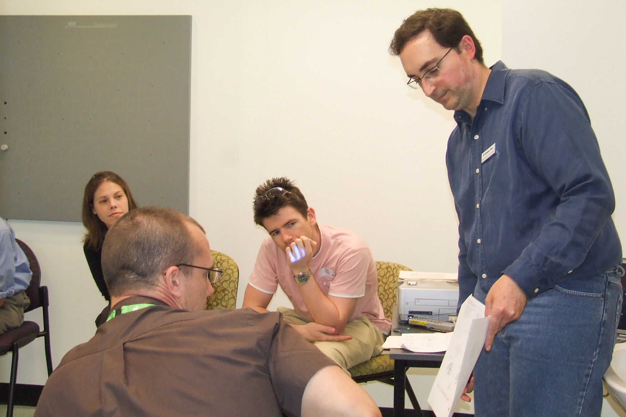 A man holding a piece of paper stands talking to a group of people sitting in chairs in a room with a notice board on the wall