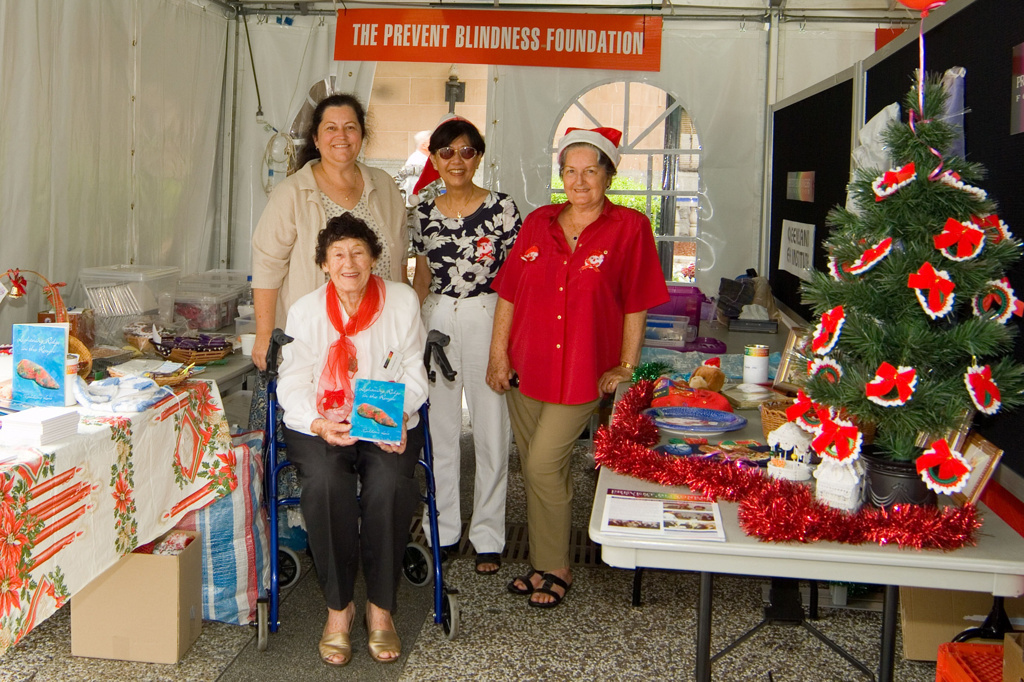 Four women are in a marquee with tables covered in Christmas decorations and items for sale