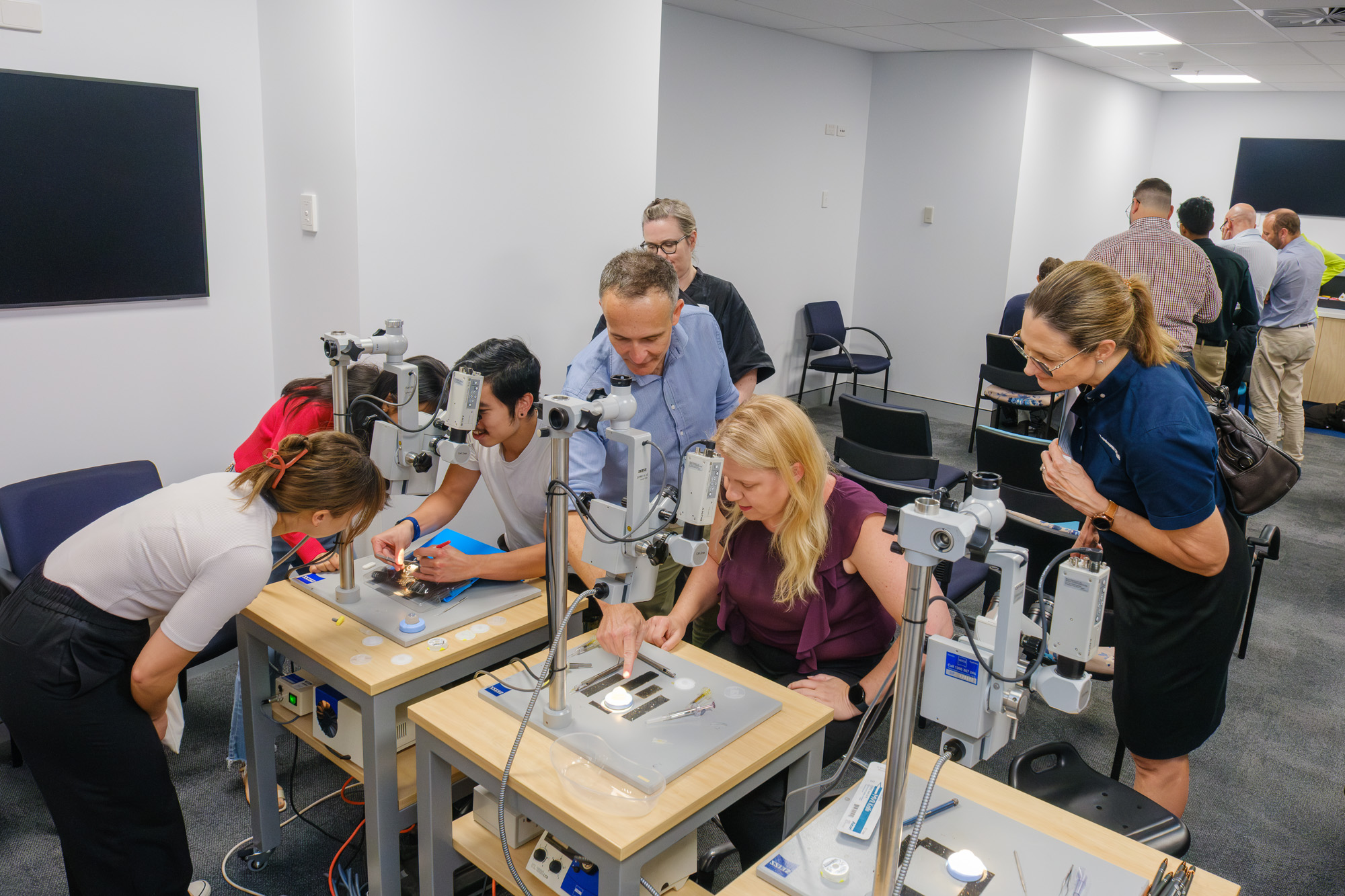 A man stands over a group of people looking through microscopes, pointing to the equipment and talking