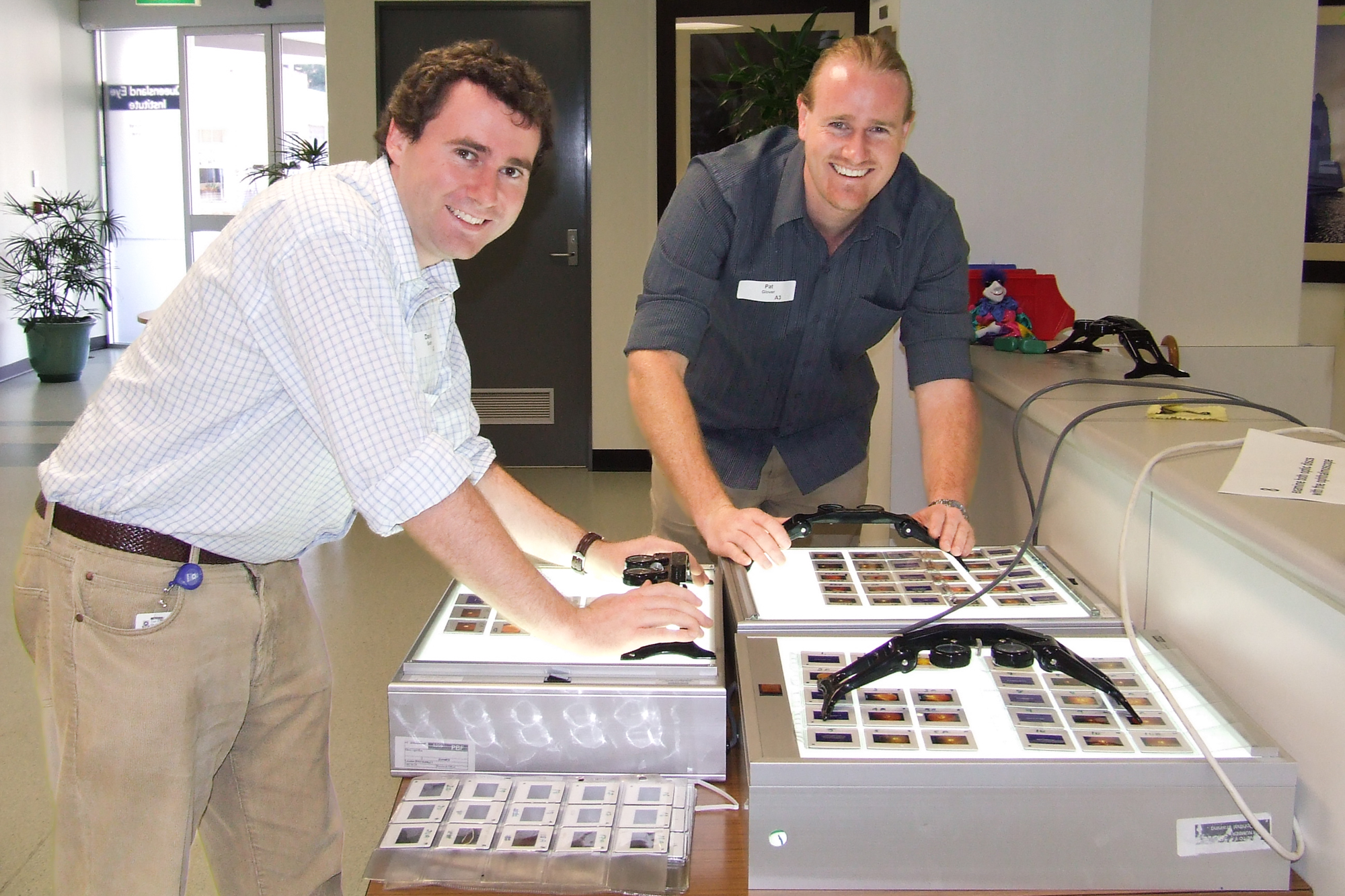 Two men smile at the camera while leaning over a box with a clear surface, lit from within, to look at slides