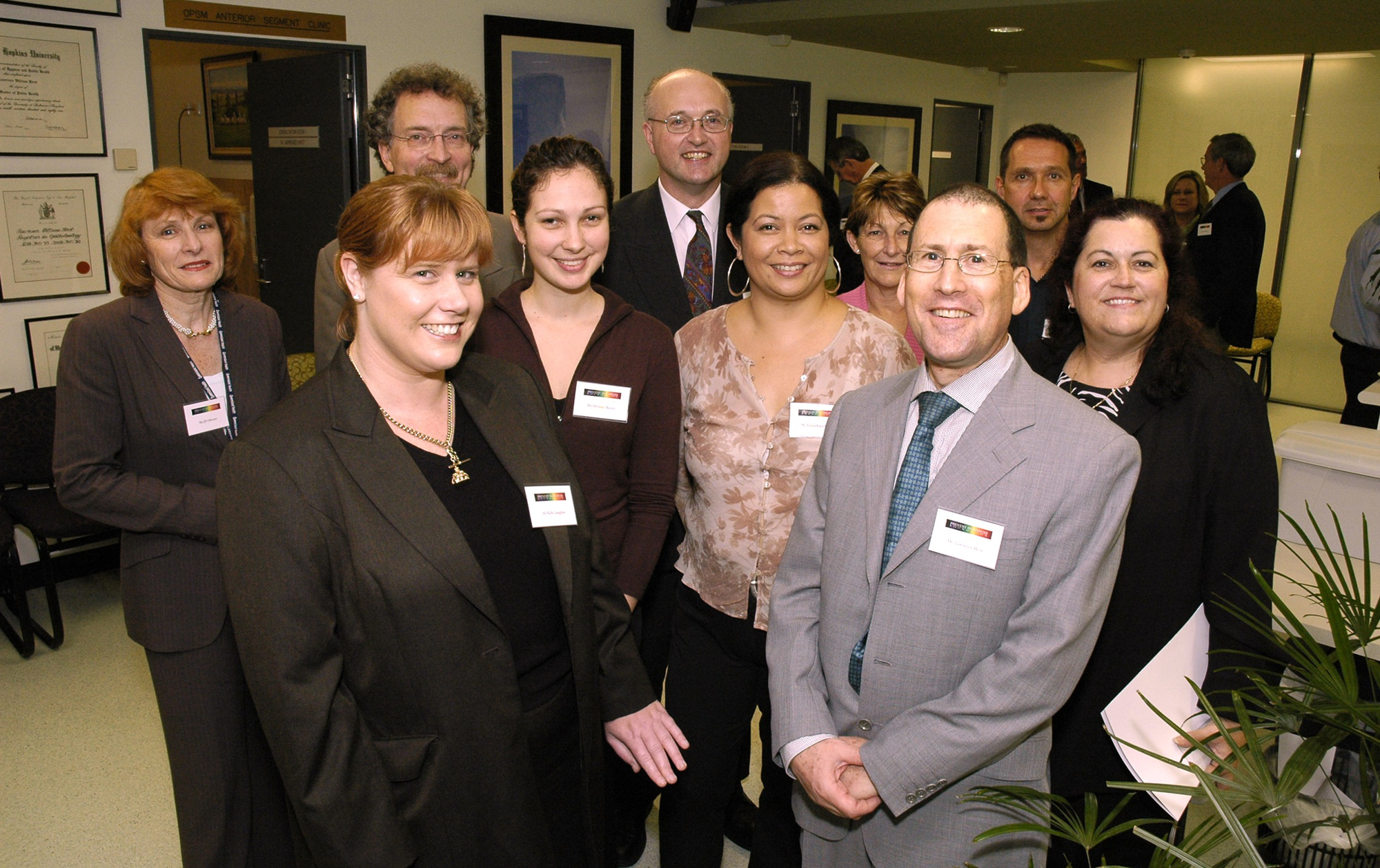 A group of men and women in suits with name tags inside a crowded room smile at the camera