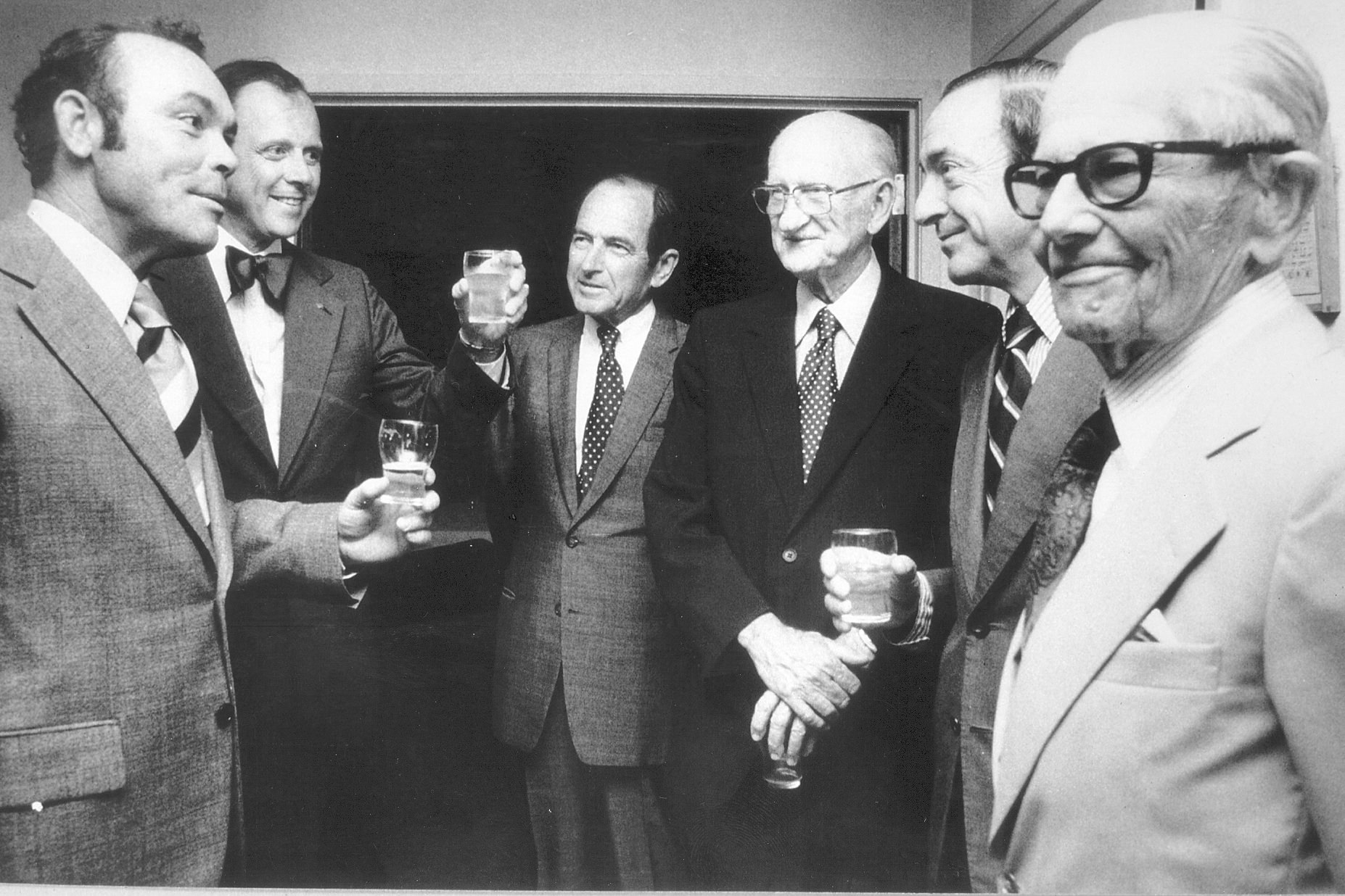 Old black and white photo of a group of men in suits talking to each other, some raising glasses in celebration