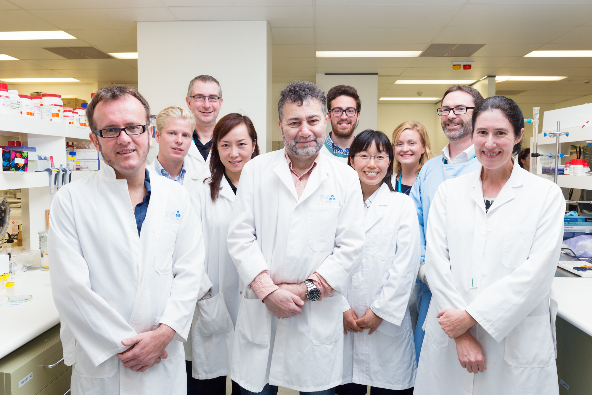 Group of 10 men and women in short white coats, standing with their hands folded in front of them in a laboratory.