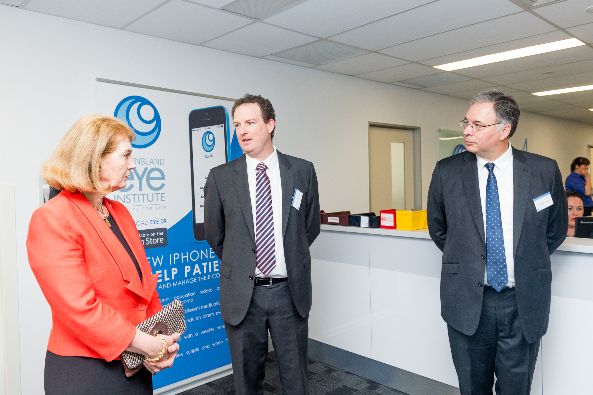 A woman in a red jacket talks to two men, standing in front of a banner advertising the Queensland Eye Institute