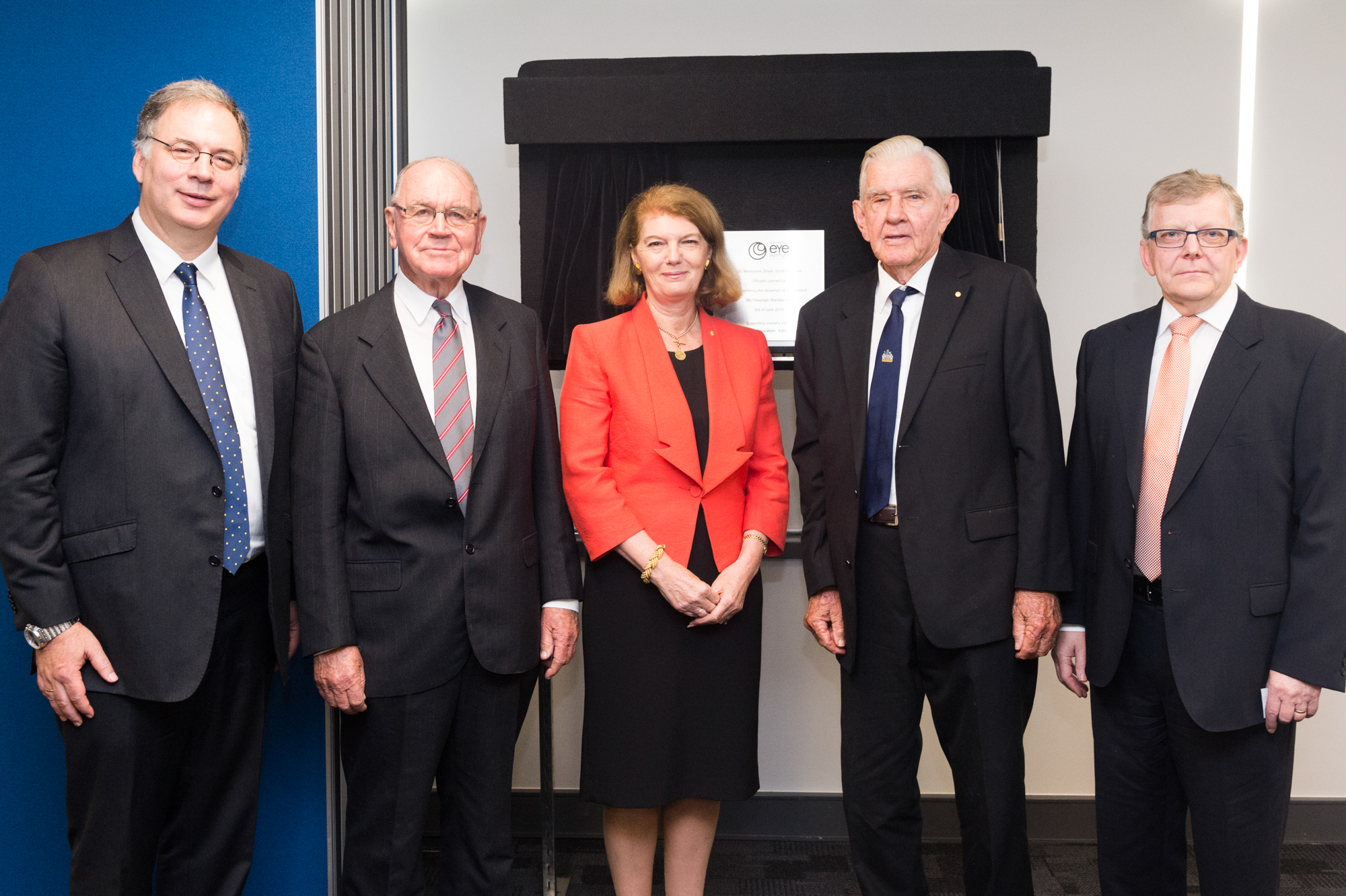 A woman in a red jacket stands in the centre with four men in dark suits beside her. They stand in front of brass plaque, mounted on a display board.