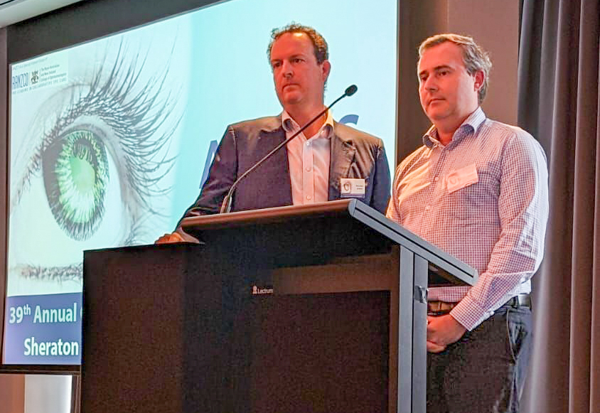 Two men speak from behind a lectern. Behind them is a large screen with the image of an eye on it.