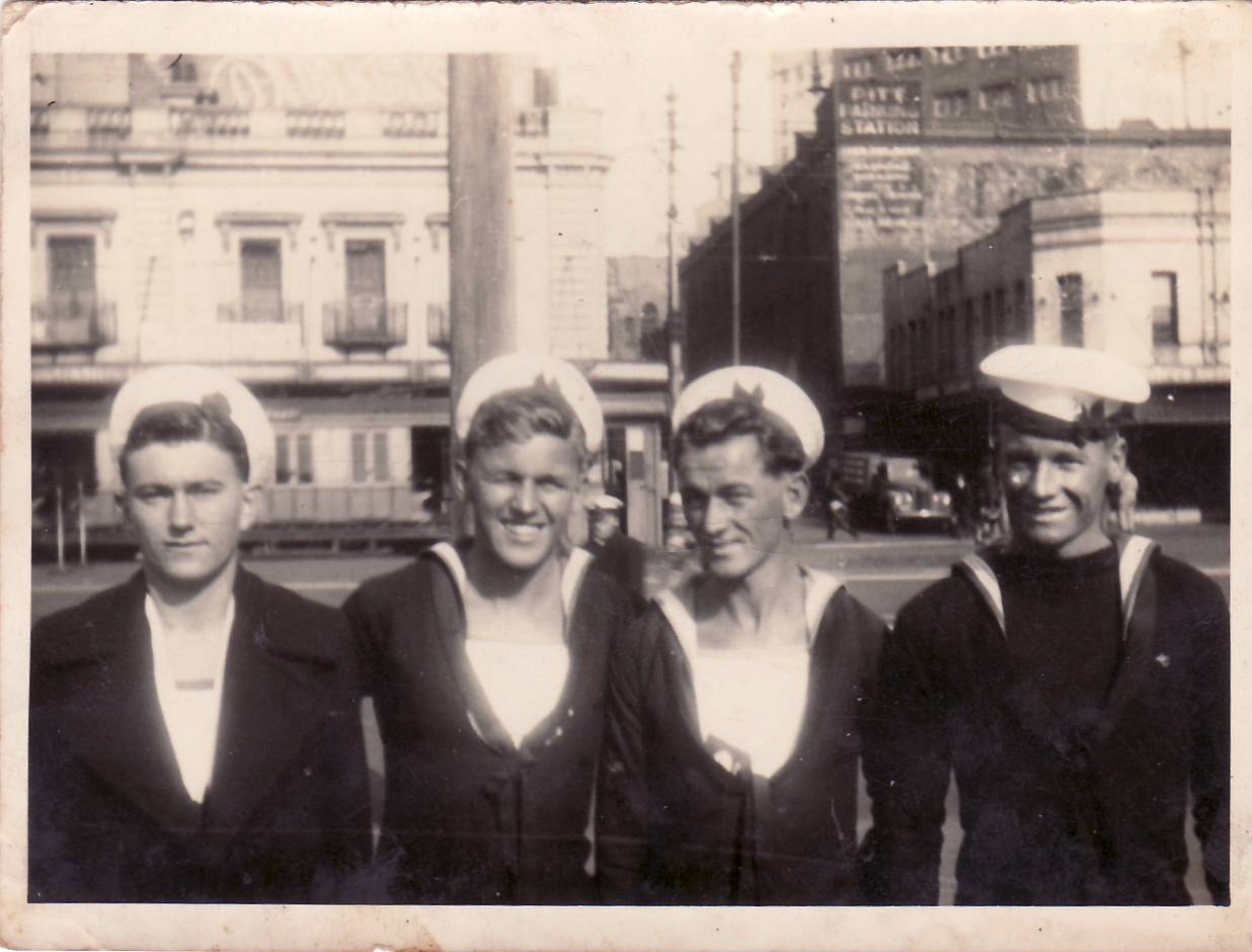 Old black and white photo of four sailors standing arm-in-arm with buildings and traffic behind them