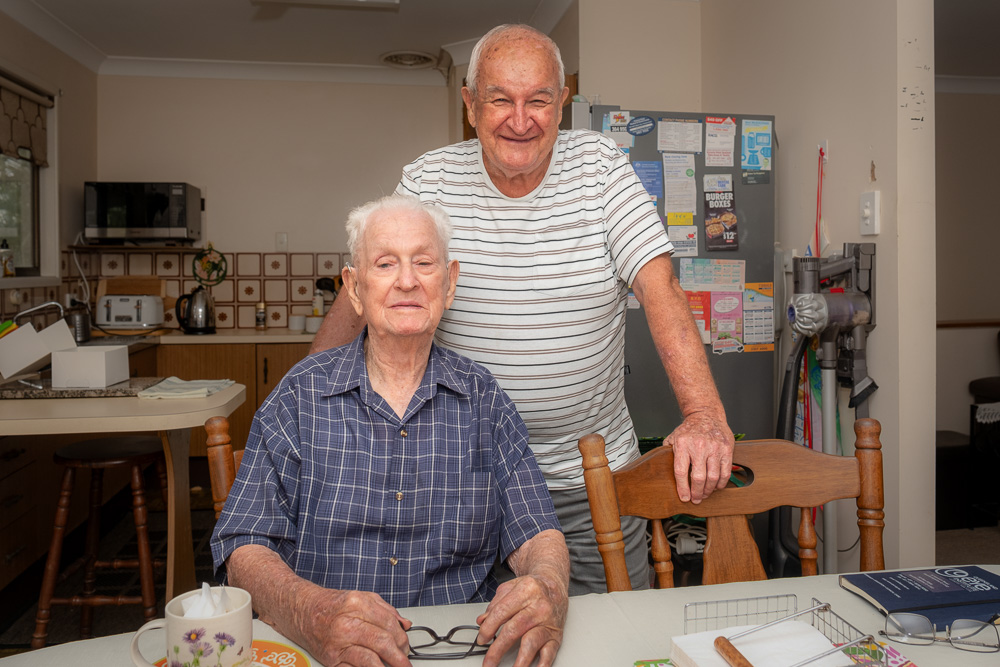 An elderly man is seated at a kitchen table, with another man standing behind him. There is a cup and saucer and napkins on the table in front of them. The elderly man holds a pair of spectacles in his hands.