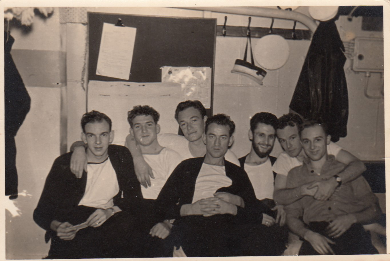 Old black and white photo of seven young men with their arms around each other, sitting on a sofa below deck on a ship.