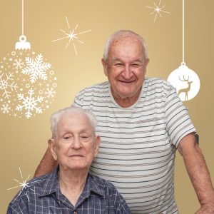 Close up image of two men, one seated and one standing. The image has a gold background with white Christmas decorations.