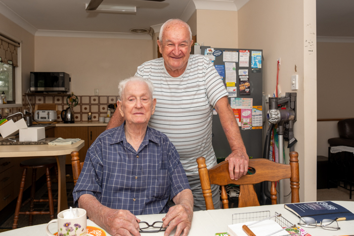 Two men at a dining table with a kitchen behind them. One is seated and the other stands.