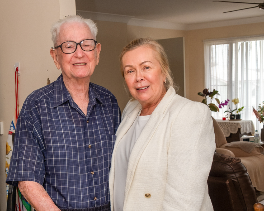 An older man in a blue checked shirt with dark rimmed glasses stands beside a woman with blonde hair wearing a white blazer