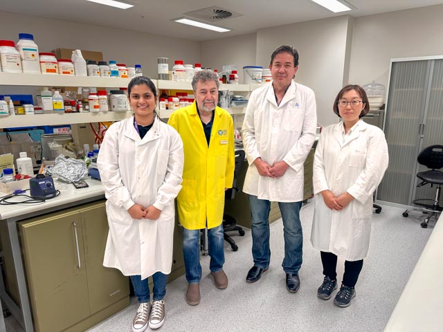 Two men and two women wearing lab coats stand side by side in front of a laboratory bench and shelves holding chemicals.