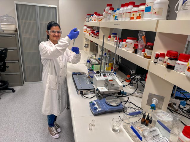 A young woman with long brown hair wears a white lab coat, safety goggles and blue rubber gloves. She stands at a laboratory bench holding an eye dropper and a beaker.