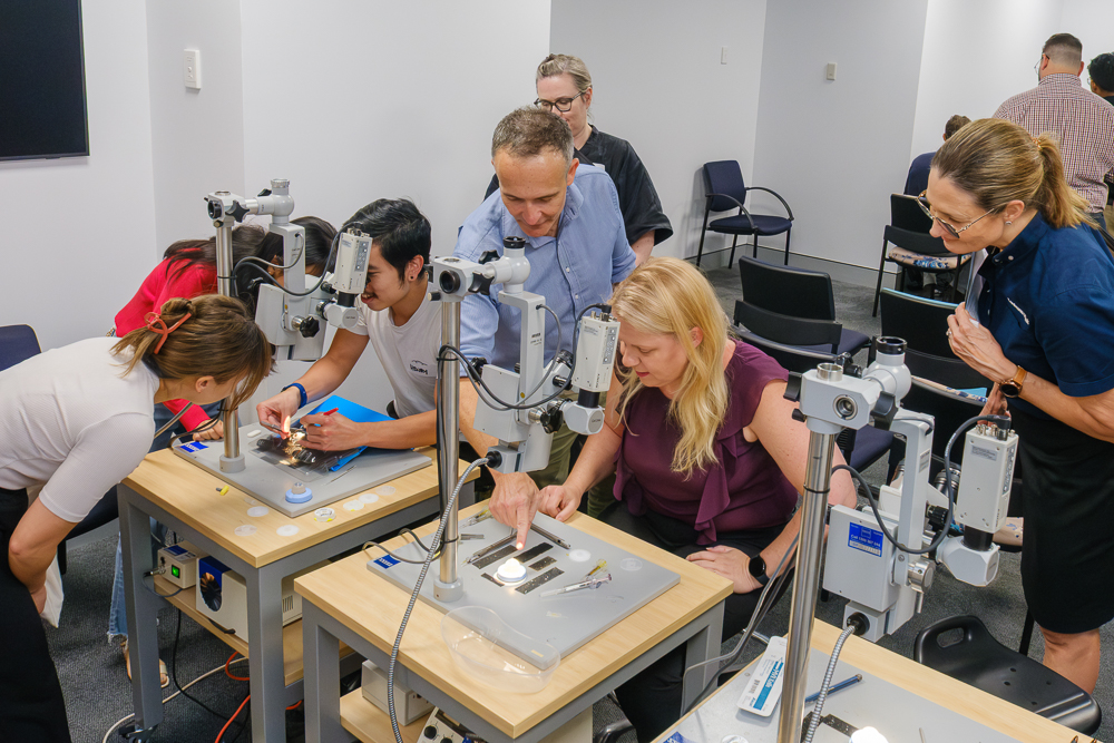 A man stands beside a woman who is looking through a microscope. The man is talking and pointing to the microscope. Another man and woman stand nearby, looking on.