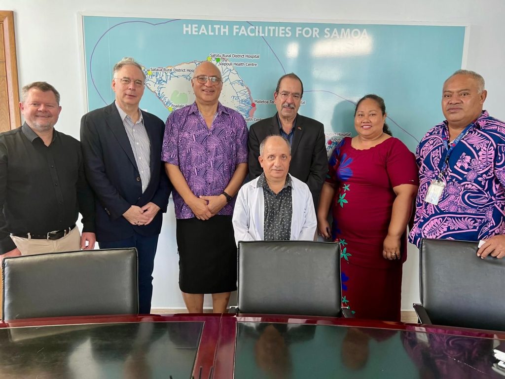 Six men and one woman stand side-by-side behind a conference table and chairs. On the wall behind them is a map of Samoa.