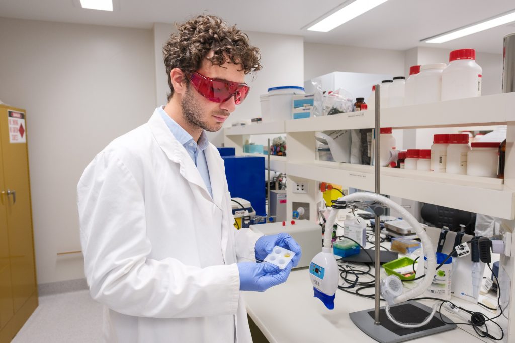 A young man with curly hair wearing safety goggles and a lab coat holds a small tray containing four samples of a gel substance.