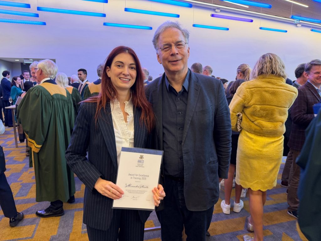 A woman in a pinstriped suit holds a certificate and stands beside a man. They are in a crowded room and are both smiling.