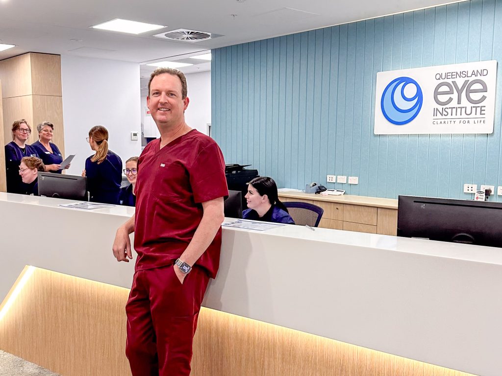 A man in maroon medical scrubs leans against a reception counter. Behind the counter some women in navy medical scrubs are talking or working at computer screens.