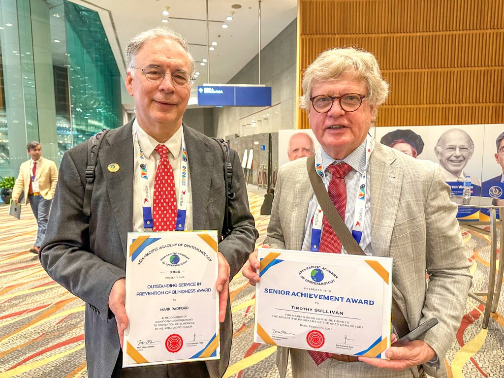 Two men in suits wearing lanyards around their necks stand side by side in the foyer of a large conference centre. They both hold certificates and are smiling at the camera.