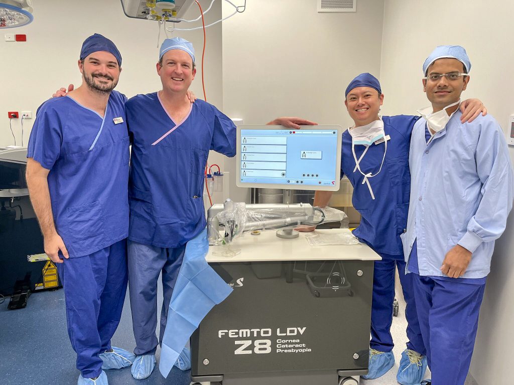 Four men in blue hospital scrubs stand beside a computer monitor and medical laser