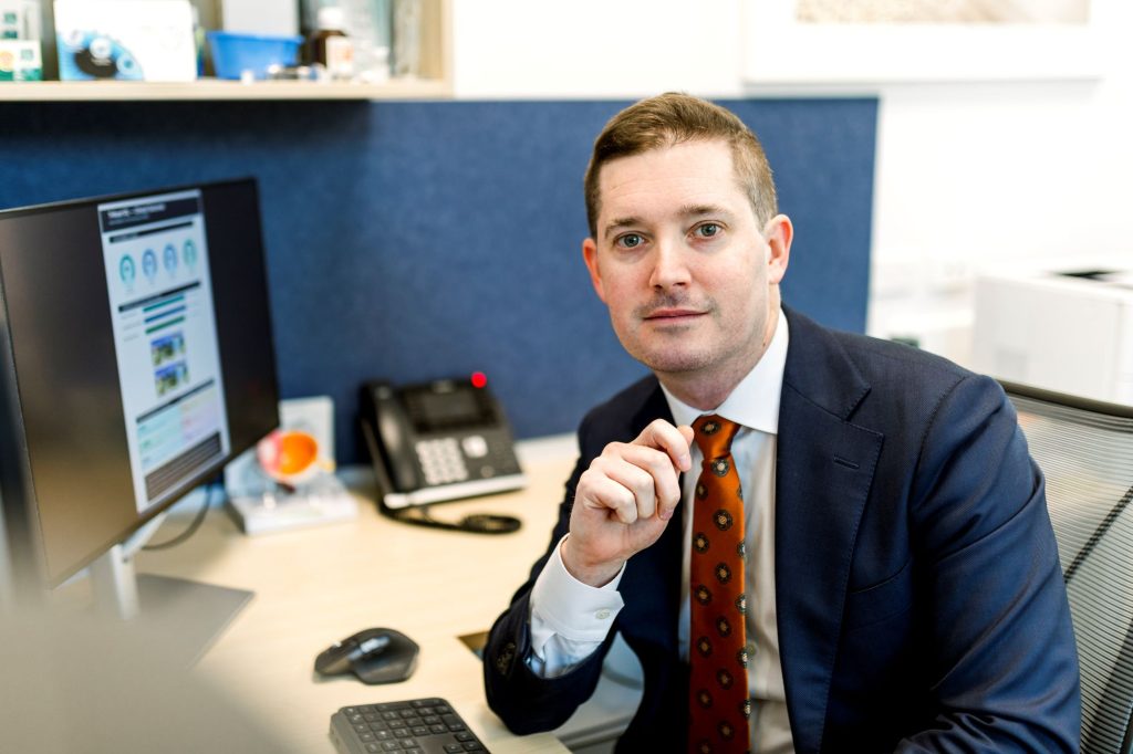 Man in a blue suit and red tie sits at a desk with a computer behind him