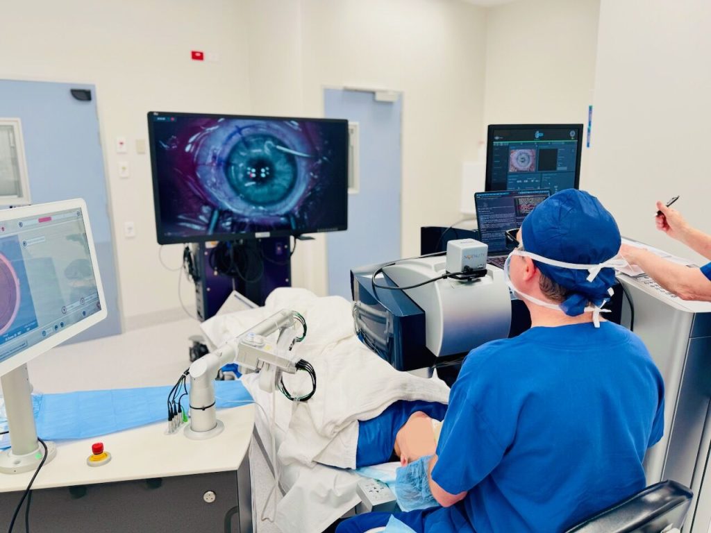 An ophthalmologist is seated and performing eye surgery on a patient while looking at a screen