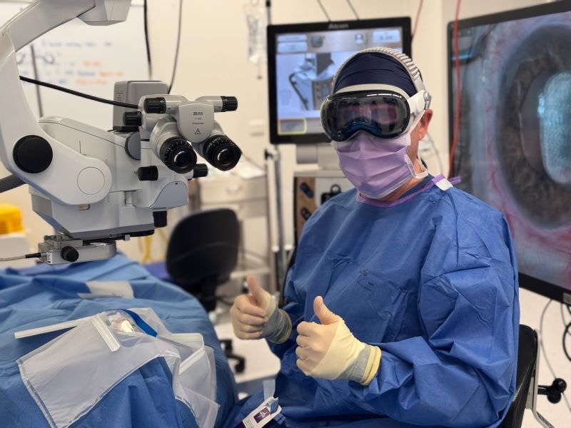 A surgeon in a mask and blue theatre gown wears a headset with goggles beside a surgical microscope and monitor screen.