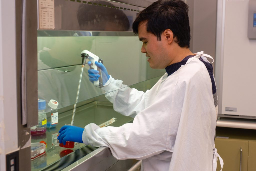 A young man with short dark hair wearing a white lab coat and blue rubber gloves is working with scientific instruments in a sterile space